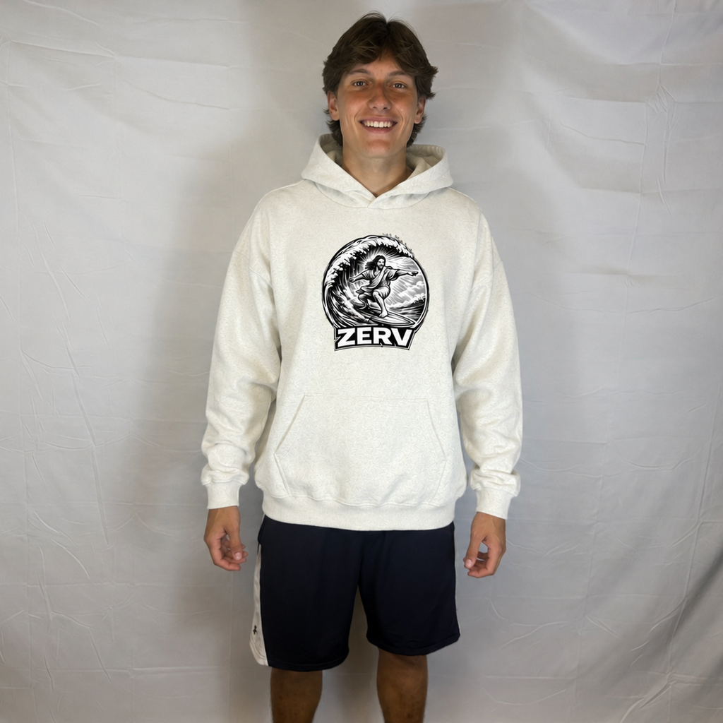 A man modeling a cream-colored hoodie with a black circular graphic on the chest featuring a surfer and the word 'ZERV'. He is wearing dark navy shorts and standing in front of a wrinkled white fabric backdrop.