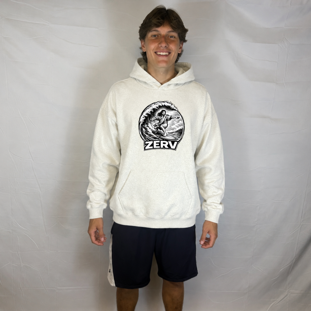 A man modeling a cream-colored hoodie with a black circular graphic on the chest featuring a surfer and the word 'ZERV'. He is wearing dark navy shorts and standing in front of a wrinkled white fabric backdrop.